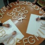 Holzbuchstaben im Kreis auf einem Tisch, rundherum Menschen