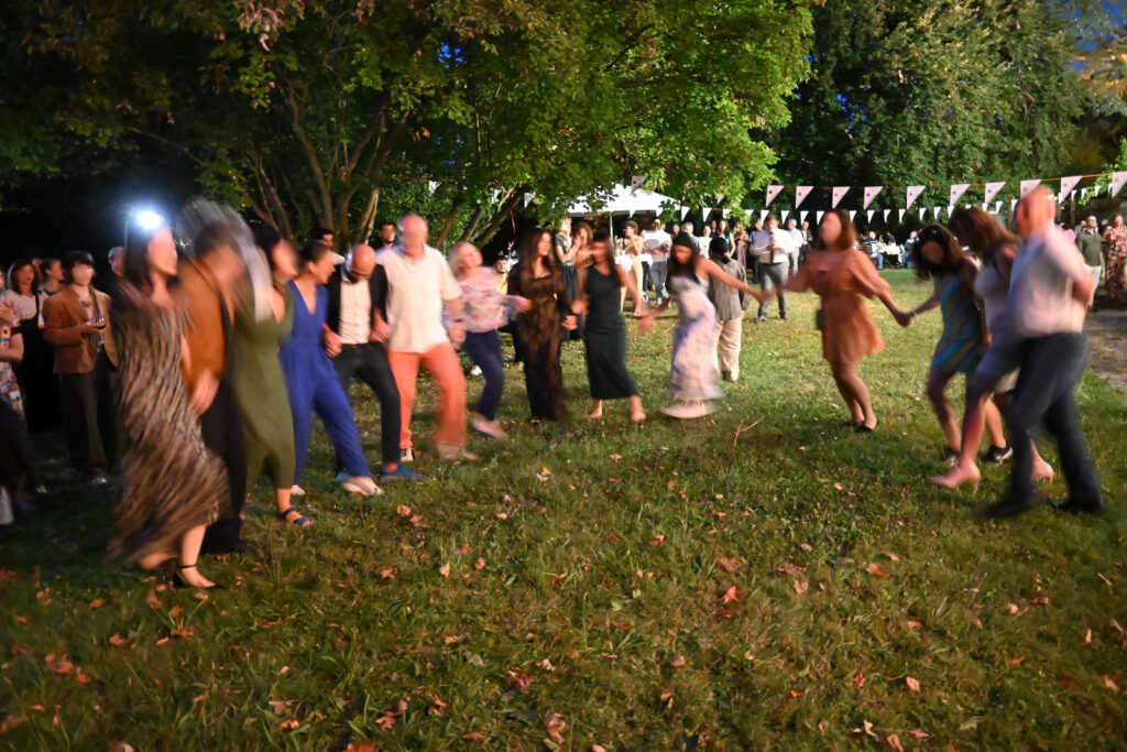 Mehrere Frauen und Männer tanzen und halten sich dabei an den Händen. Sie sind in einem Garten. Im Hintergrund sieht man einen großen Busch und Bäume, Girlanden, einen Pavillon und weitere Menschen, die teilweise bei Stehtischen stehen, ein Weinglas halten oder auf Gartenstühlen zusammensitzen.