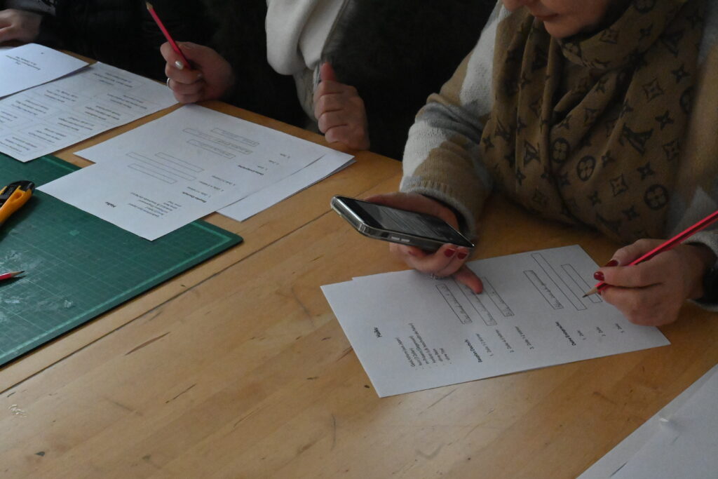 Man sieht die Fläche des Arbeitstisches. Darauf sieht man eine Arbeitsunterlage, einige Arbeitsblätter. Am oberen Rand des Bildes sieht man die Hände derer Teilnehmerinnen, die gerade die Arbeitsblätter bearbeiten. Eine Teilnehmerin hält ihr Smartphone in der einen Hand, einen Bleistift in der anderen.