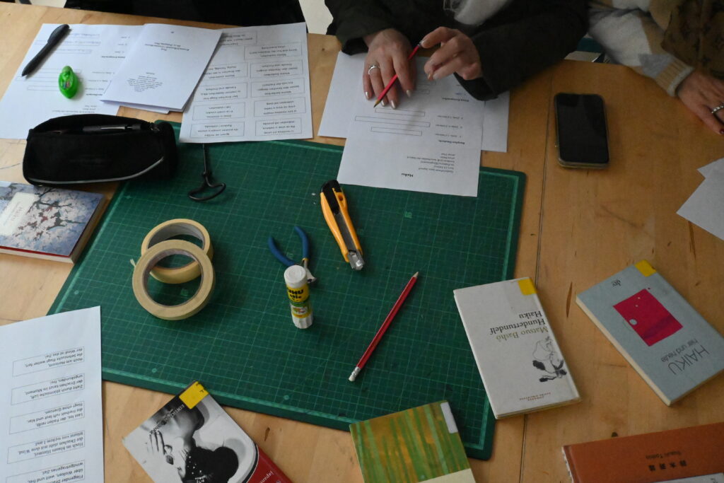 Vogelperspektive auf einen der Basteltische. Man sieht eine Bastelunterlage, mehrere Klebebänder, einen UHU-Stick, ein Stanley-Messer, einen Bleistift, Arbeitsblätter und einige Bücher mit Gedichten auf dem Tisch verteilt. Am oberen Rand des Bildes sieht man die Hände zweier Kursteilnehmerinnen, eine hält einen Bleistift.