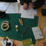 Vogelperspektive auf einen der Basteltische. Man sieht eine Bastelunterlage, mehrere Klebebänder, einen UHU-Stick, ein Stanley-Messer, einen Bleistift, Arbeitsblätter und einige Bücher mit Gedichten auf dem Tisch verteilt. Am oberen Rand des Bildes sieht man die Hände zweier Kursteilnehmerinnen, eine hält einen Bleistift.