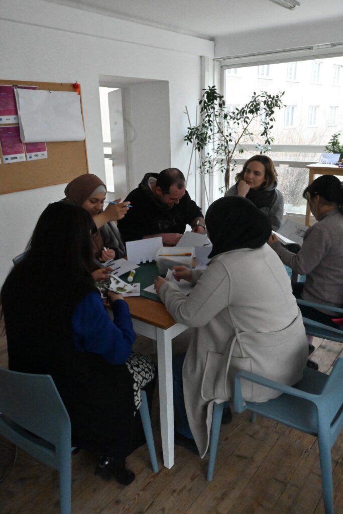 Mehrere Kursteilnehmer*innen und eine UKI-Kursleiterin sitzen rund um den Basteltisch. Auf dem Tisch und in den Händen der Personen sind gebastelte Billets, Scheren und Stifte.