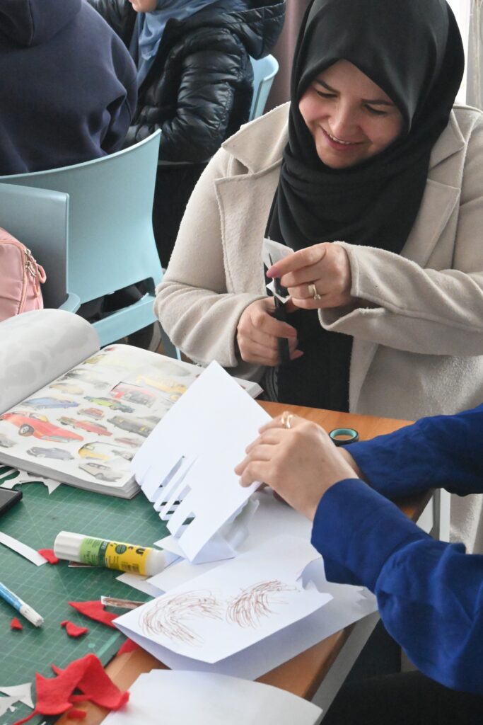 Eine Teilnehmerin sitzt am Basteltisch. Vor ihr am Tisch liegen Filz- und Papierschnipsel, Kleber und ein Stift. Die Teilnehmerin lächelt, während sie ein auf Papier gedrucktes Bild ausschneidet. Rechts im Bild sieht man die Hände einer weiteren Teilnehmerin, die gerade bastelt. Weiter links im Bild, sieht man ein geöffnetes Heft. Auf den sichtbaren Seiten des Heftes sind ausgeschnittene Bilder von unterschiedlichen Fahrzeugen aufgeklebt.