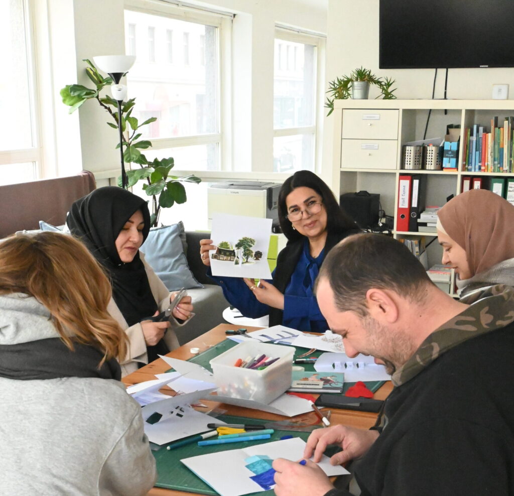 Mehrere Kurs-Teilnehmende sitzen gemeinsam an einem Tisch. Sie malen, schneiden, basteln. Dabei unterhalten sie sich und lächeln. Eine Teilnehmerin lächelt stolz in die Kamera und hält ihre selbstgebasteltes Billet hoch.