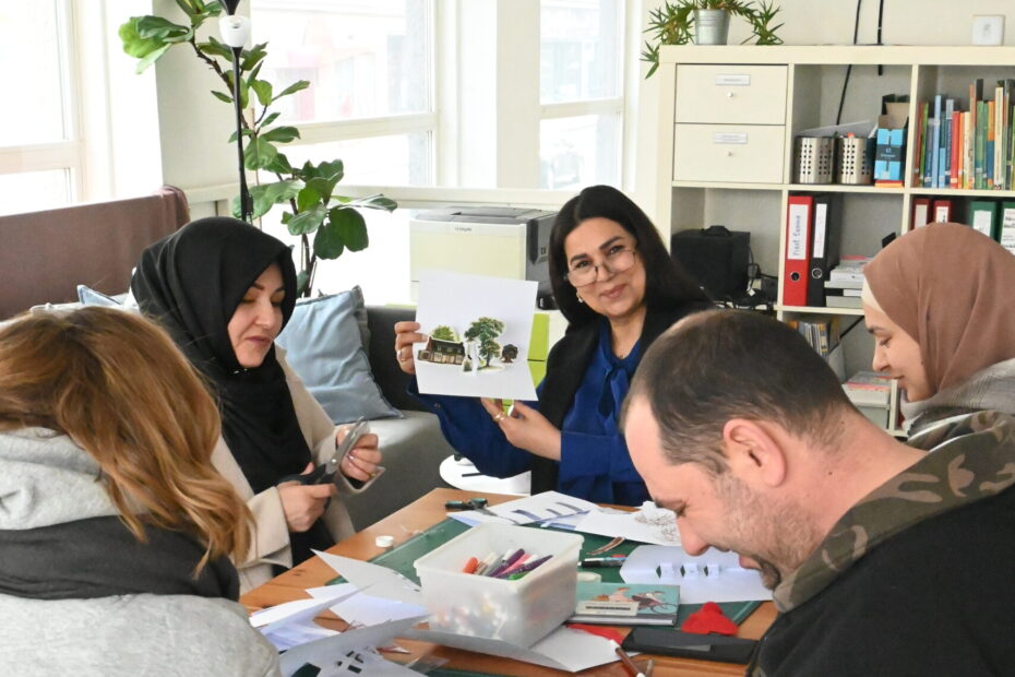 Mehrere Kurs-Teilnehmende sitzen gemeinsam an einem Tisch. Sie malen, schneiden, basteln. Dabei unterhalten sie sich und lächeln. Eine Teilnehmerin lächelt stolz in die Kamera und hält ihre selbstgebasteltes Billet hoch.