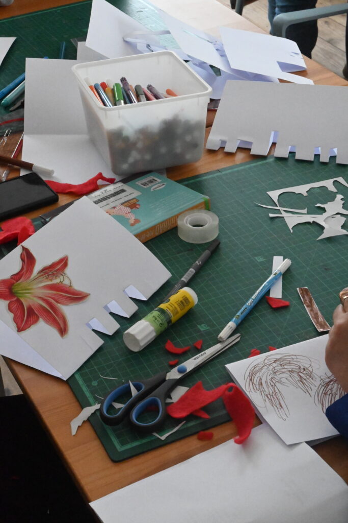 Stifte, Klebstoff, eine Schere und Papierschnipsel liegen auf dem Arbeitstisch sowie ein Billet, das von einer Teilnehmerin gestaltet wurde. Darauf ist das Bild einer Blume aufgeklebt, das aus einer Zeitschrift ausgeschnitten wurde.