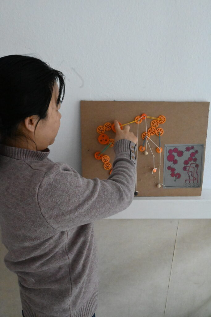 Eine Kursteilnehmerin steht seitlich, leicht mit dem Rücken zur Kamera gewandt. Sie ist zur Wand hinter sich gewandt. Dort hängt ein Pappkarton mit einem Gebilde aus kleinen Zahnrädern, die durch Fäden verbunden sind. Das Gebilde erinnert an das Innere einer Maschine.