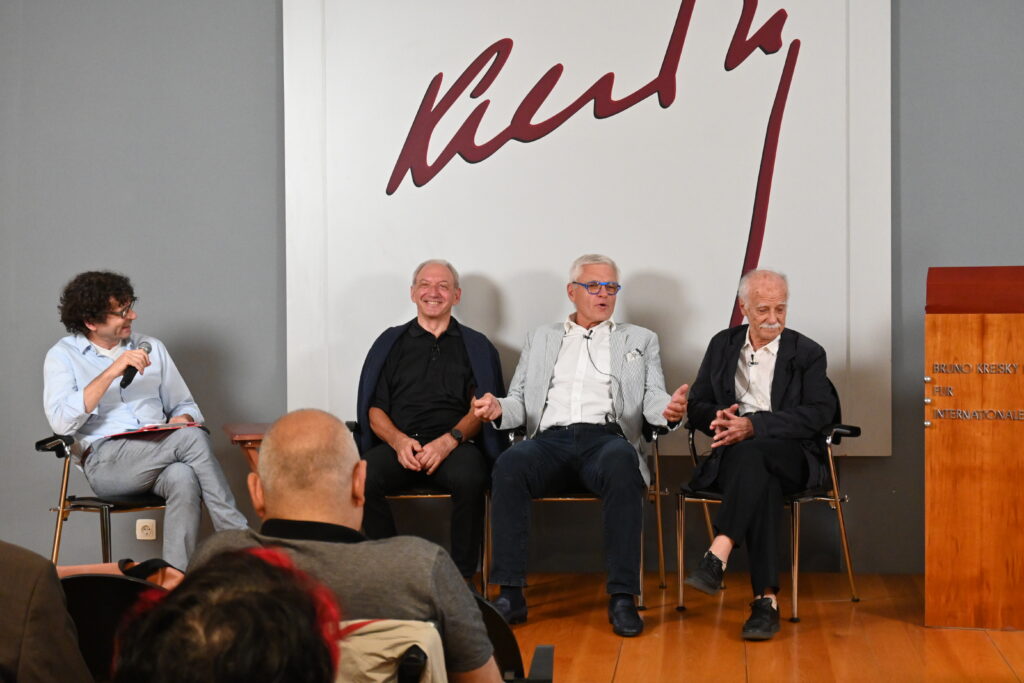 Vier Männer sitzen auf Stühlen nebeneinander auf einer kleinen Bühne. Der Mann ganz links, mittleren Alters, hält ein Mikrofon und lächelt den anderen Männern zu. Die anderen Männer sind etwas älter. Der zweite Mann grinst in das Publikum aus dessen Perspektive das Foto ist. Der dritte Mann gestikuliert und spricht gerade. Der vierte Mann hat die Beine überschlagen und die Finger verschränkt und blickt lächelnd auf den Boden. An der Wand hinter den Männern steht groß der Schriftzug "Kreisky".