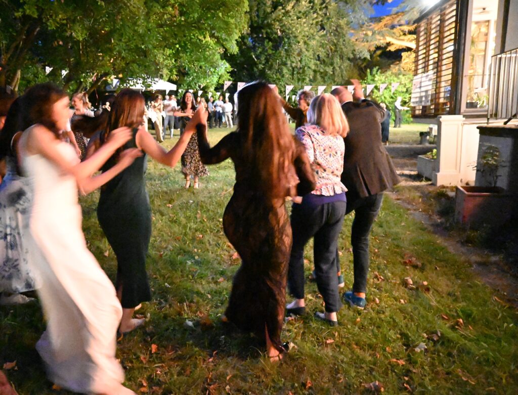 Mehrere Frauen und Männer tanzen und halten sich dabei an den Händen. Sie sind in einem Garten. Im Hintergrund sieht man Girlanden.