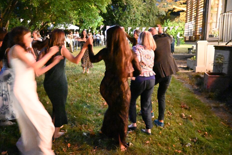 Mehrere Frauen und Männer tanzen und halten sich dabei an den Händen. Sie sind in einem Garten. Im Hintergrund sieht man Girlanden.