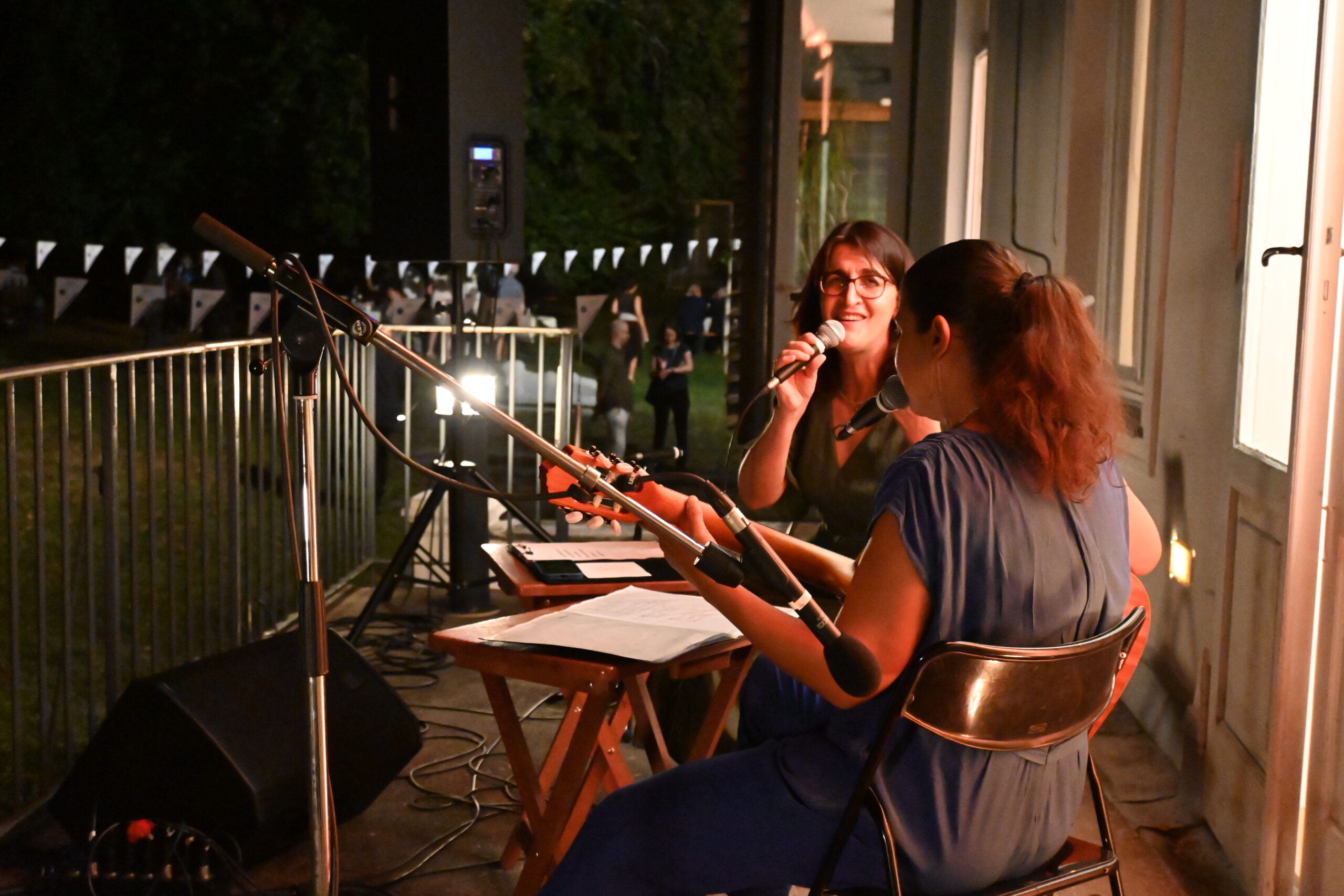 Die Musikerinnen Sakîna Teyna, Özlem Bulut sitzen auf einem niedrigen Balkon nach links bzw. richtung Garten. Teyna hält ein Mikrofon, vor und neben Bulut sind zwei Stehmikrofone. Vor den Musikerinnen zwei kleine Tischchen mit Noten und Liedtexten und zwei Verstärker. Teyna blickt richtung Kamera und lächelt während sie singt. Bulut sitzt mit dem Rücken leicht schräg zur Kamera. Im Hintergrund sieht man den Garten mit Girlanden und einigen Personen. Es ist schon dunkel. Das Licht kommt von Scheinwerfern und aus den Fenstern des Hauses, das rechts im Bild, anschließend an den Balkon zu sehen ist.
