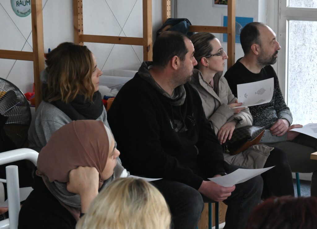 Mehrere Personen sind von der Seite zu sehen. Sie sitzen auf Stühlen und haben Zettel in der Hand. Sie scheinen, aufmerksam jemandem zuzuhören, der oder die sich außerhalb des Bildausschnitts befindet.