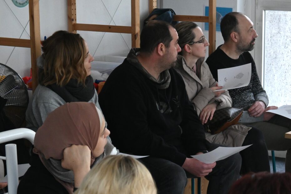Mehrere Personen sind von der Seite zu sehen. Sie sitzen auf Stühlen und haben Zettel in der Hand. Sie scheinen, aufmerksam jemandem zuzuhören, der oder die sich außerhalb des Bildausschnitts befindet.