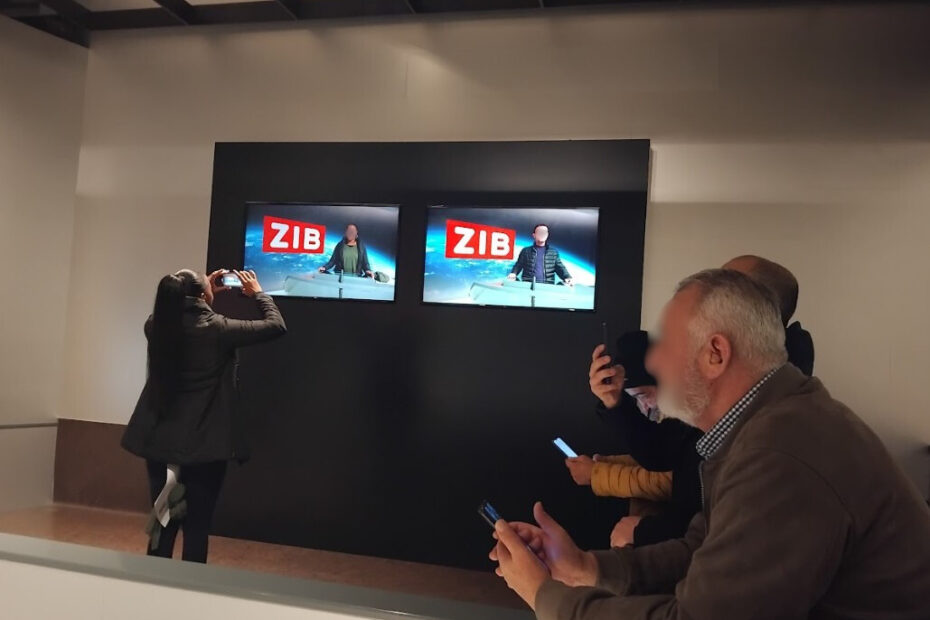 Ein nachgebildetes ZIB-Fernsehstudio. Hinten an der Wand sieht man zwei Monitore: Auf einem sieht man eine UKI-Kursteilnehmerin auf dem anderen einen UKI-Kursteilnehmer. Auf beiden Monitoren sieht im Hintergrund den typischen ZIB-Hintergrund, den man in der Nachrichtensendung immer hinter den Nachrichtensprecher*innen sieht. Rechts im Bild sieht man einige weitere UKI-Teilnehmer sitzend und mit Smartphones in den Händen, um Fotos zu machen. Direkt vor einem der Montore steht eine weitere UKI-Kursteilnehmerin mit dem Rücken zur Kamera. Sie fotografiert mit ihrem Smartphone die Monitore.