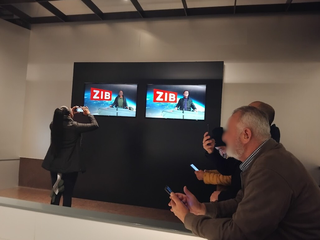 Ein nachgebildetes ZIB-Fernsehstudio. Hinten an der Wand sieht man zwei Monitore: Auf einem sieht man eine UKI-Kursteilnehmerin auf dem anderen einen UKI-Kursteilnehmer. Auf beiden Monitoren sieht im Hintergrund den typischen ZIB-Hintergrund, den man in der Nachrichtensendung immer hinter den Nachrichtensprecher*innen sieht. Rechts im Bild sieht man einige weitere UKI-Teilnehmer sitzend und mit Smartphones in den Händen, um Fotos zu machen. Direkt vor einem der Montore steht eine weitere UKI-Kursteilnehmerin mit dem Rücken zur Kamera. Sie fotografiert mit ihrem Smartphone die Monitore.