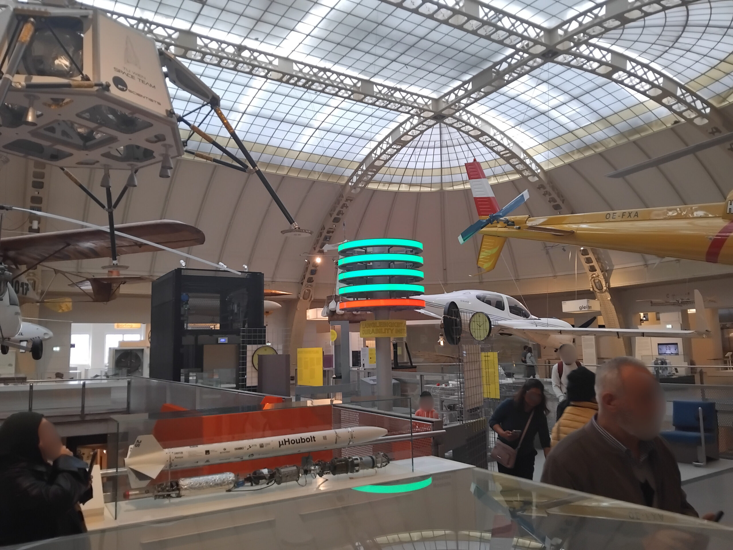 Man sieht die hohen Decken in einer großen Ausstellungshalle des Technischen Museums. Von der Decke hängen nachgebildete Modelle von kleinen, älteren Flugzeugmodellen. Einige Museumsbesucher*innen bewegen sich durch die Halle.