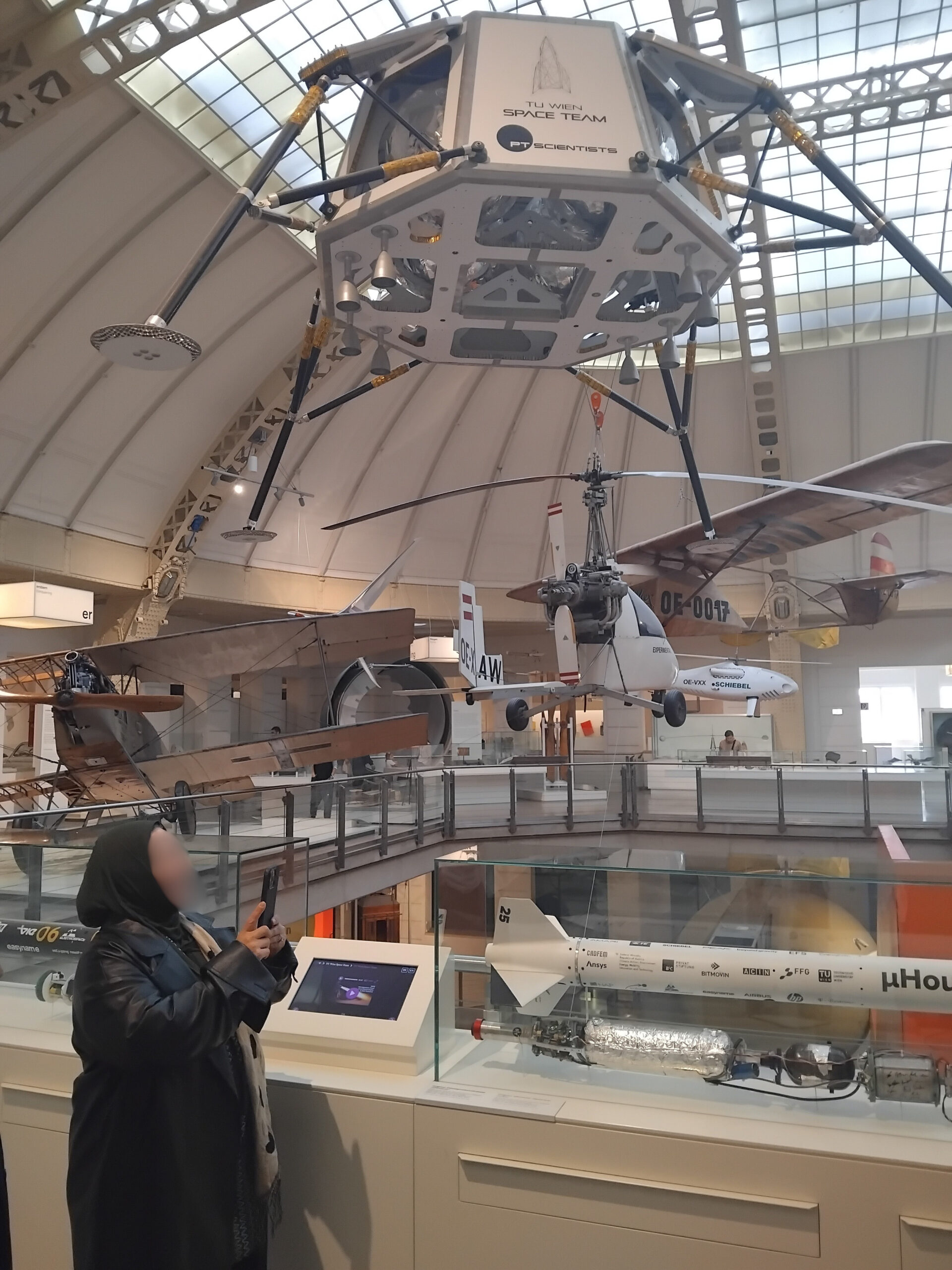 Eine große Ausstellungshalle des Technischen Museums. Man sieht von der Decke Nachbildung kleinerer Flugzeugmodelle sowie eine Nachbildung einer Rakete hängen. Eine Besucherin betrachtet die Modelle und hält ihr Smartphone zum Fotografieren bereit.