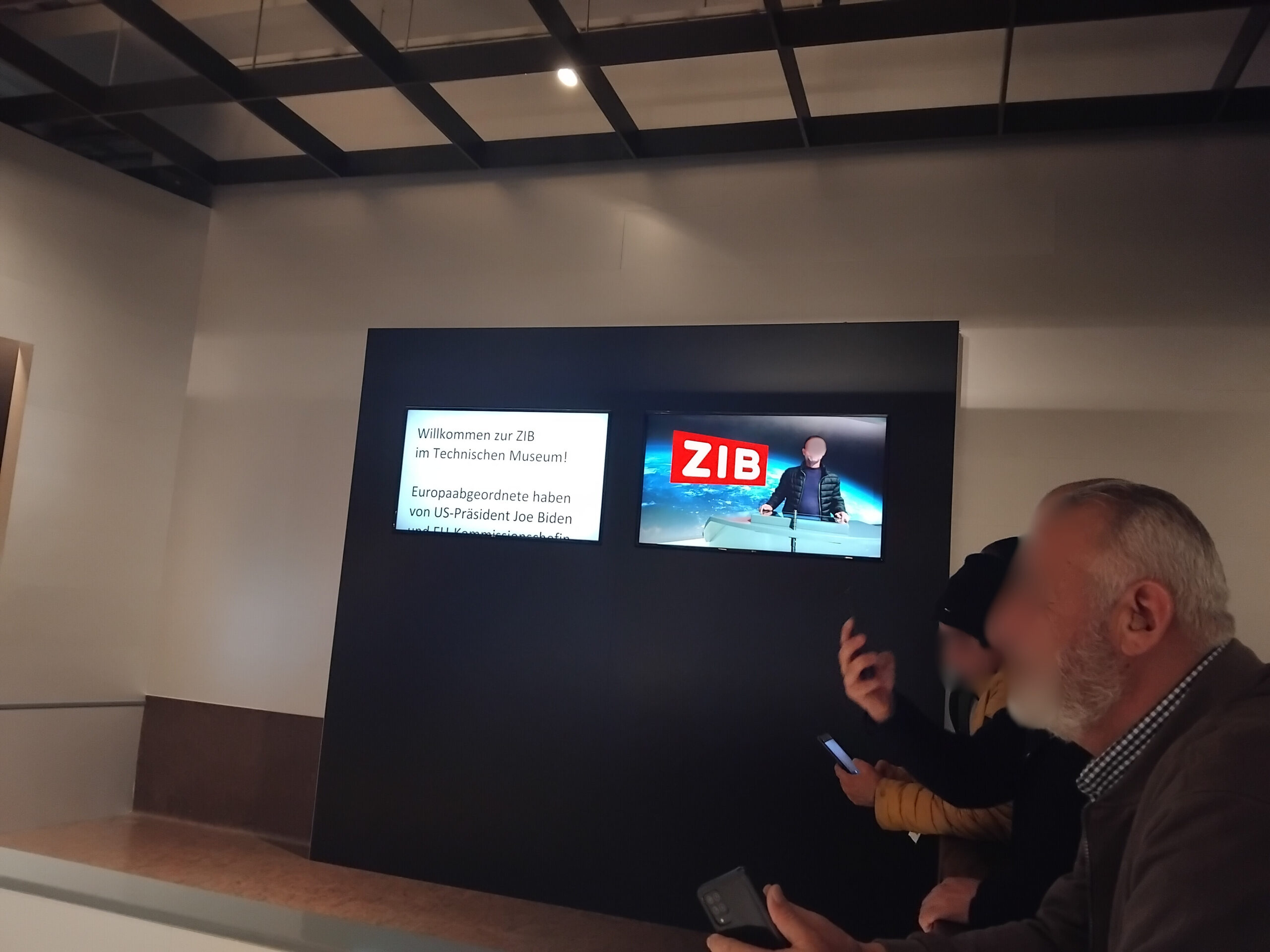 Ein nachgebildetes ZIB-Fernsehstudio. Hinten an der Wand sieht man zwei Monitore: Auf einem sieht man den Text eines Teleprompters "Willkommen zur ZIB im Technischen Museum! Europaabgeordnete haben von US-Präsident Joe Biden ...". Am anderen Bildschirm sieht man einen UKI-Kursteilnehmer mit dem typischen ZIB-Hintergrund, den man in der Nachrichtensendung immer hinter den Nachrichtensprecher*innen sieht. Rechts im Bild sieht man einige weitere UKI-Teilnehmer sitzend und mit Smartphones in den Händen, um Fotos zu machen.