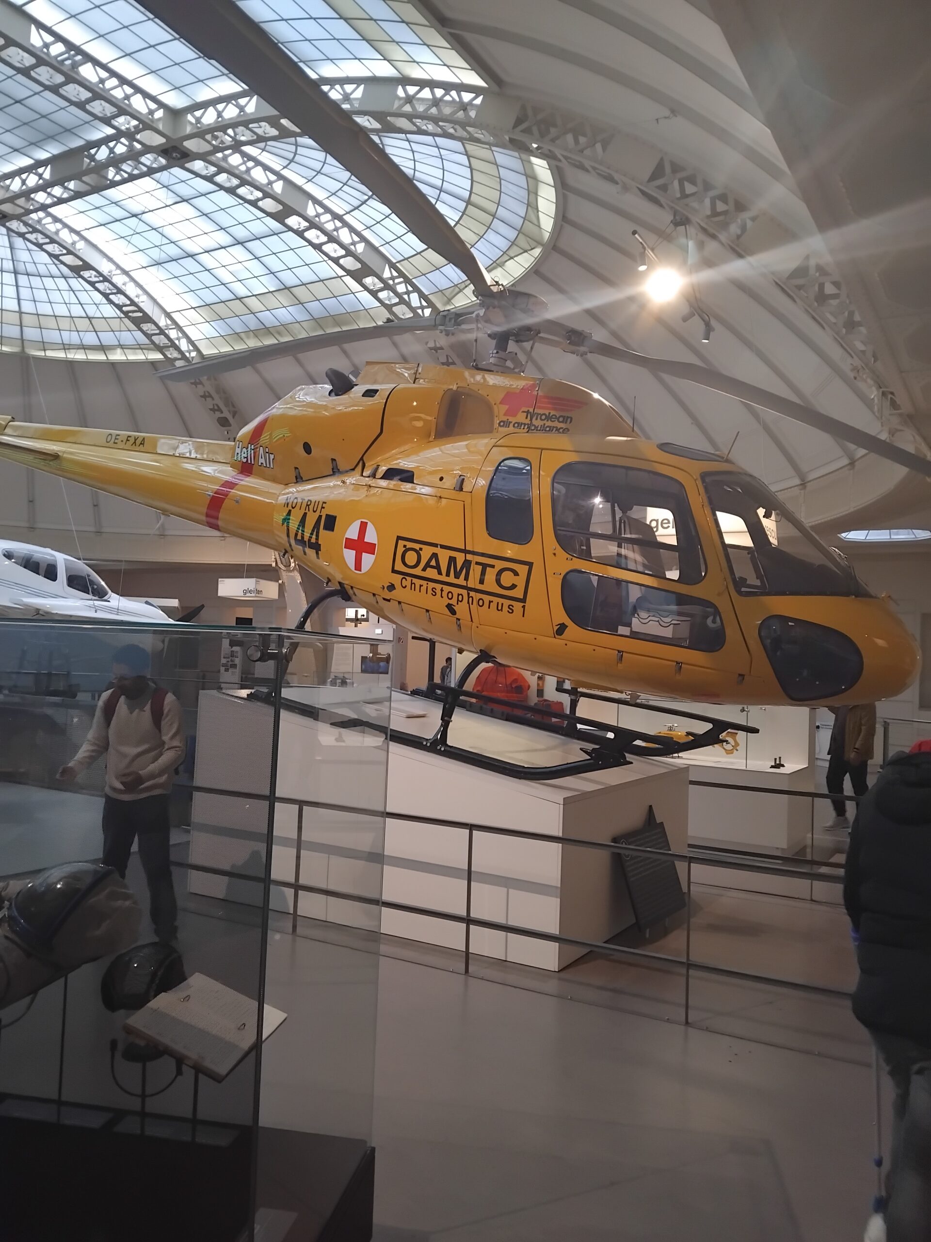 Eine große Ausstellungshalle des Technischen Museums. Von der Decke hängen nachgebaute kleine Flugzeug- und Hubschraubermodelle. Zentral im Bild ist ein gelber ÖAMTC-Hubschrauber. Einige Museumsbesucher*innen bewegen sich durch die Halle und betrachten die Ausstellungsstücke.